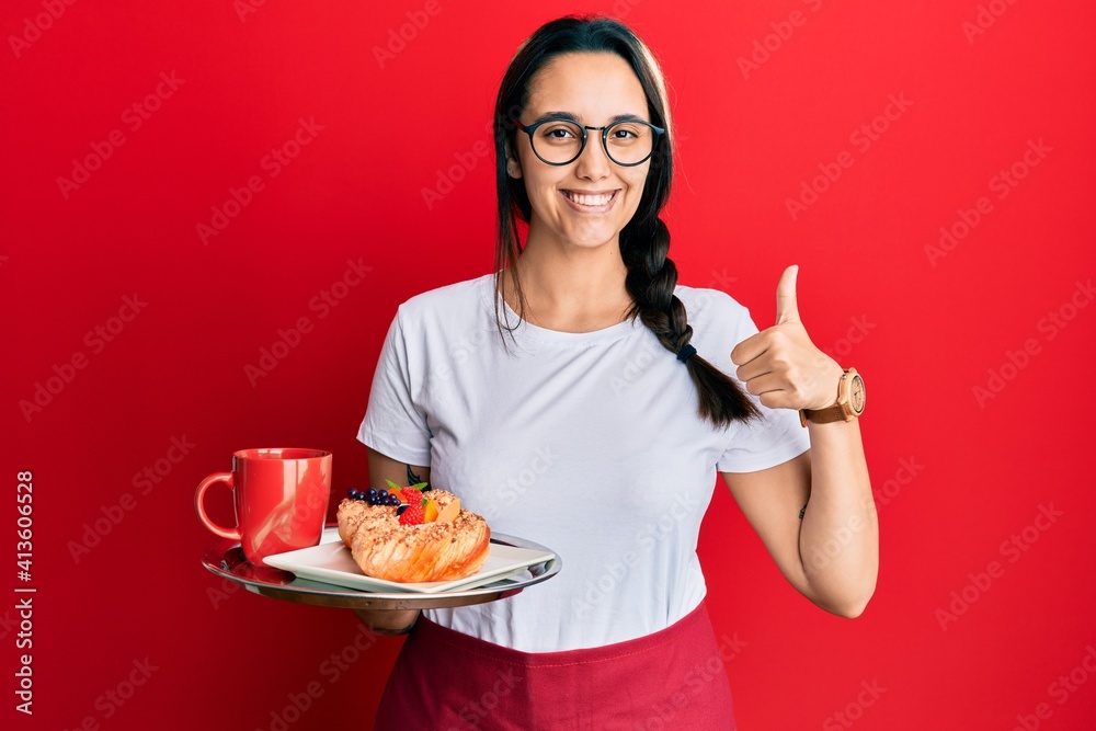Young hispanic woman wearing waitress apron holding tray with breakfast smiling happy and positive, thumb up doing excellent and approval sign