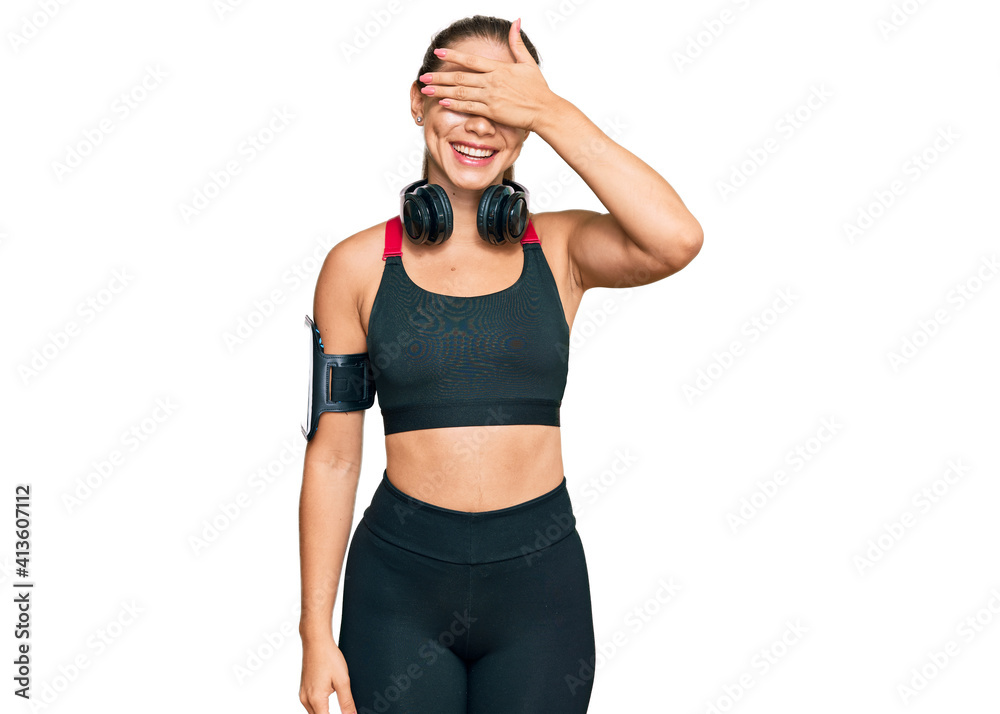 Beautiful blonde woman wearing gym clothes and using headphones smiling and laughing with hand on face covering eyes for surprise. blind concept. Beautiful blonde woman wearing gym clothes and using headphones smiling and laughing with hand on face covering eyes for surprise. blind concept.