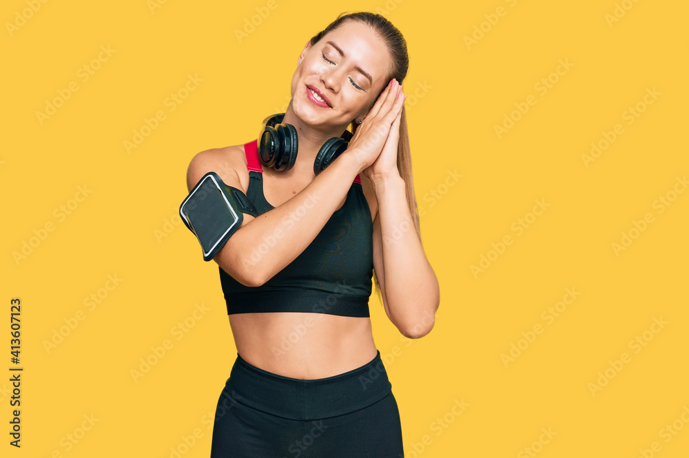 Beautiful blonde woman wearing gym clothes and using headphones sleeping tired dreaming and posing with hands together while smiling with closed eyes. Beautiful blonde woman wearing gym clothes and using headphones sleeping tired dreaming and posing with hands together while smiling with closed eyes.