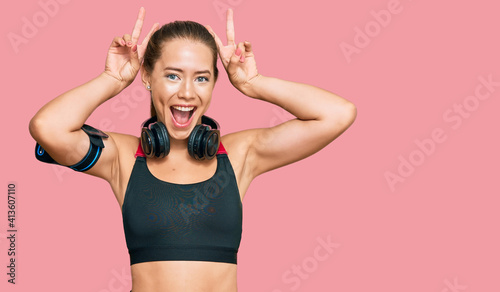Beautiful blonde woman wearing gym clothes and using headphones posing funny and crazy with fingers on head as bunny ears, smiling cheerful © Krakenimages.com