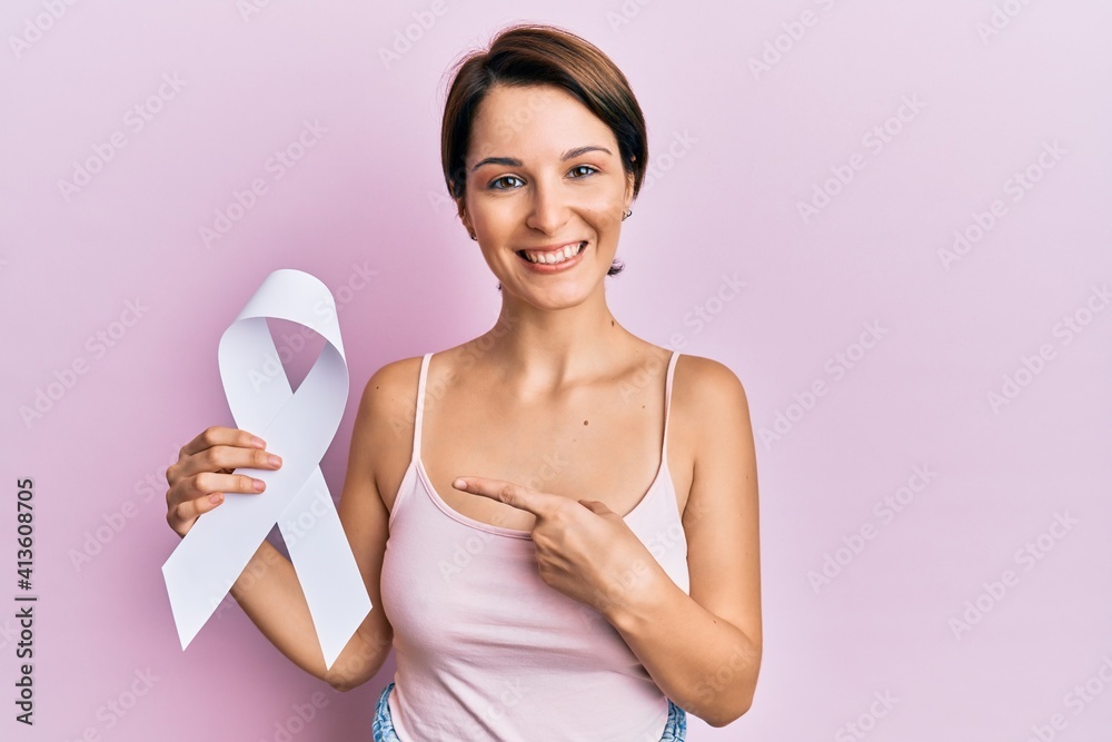 Young brunette woman with short hair holding cancer awareness white ribbon smiling happy pointing with hand and finger Young brunette woman with short hair holding cancer awareness white ribbon smiling happy pointing with hand and finger