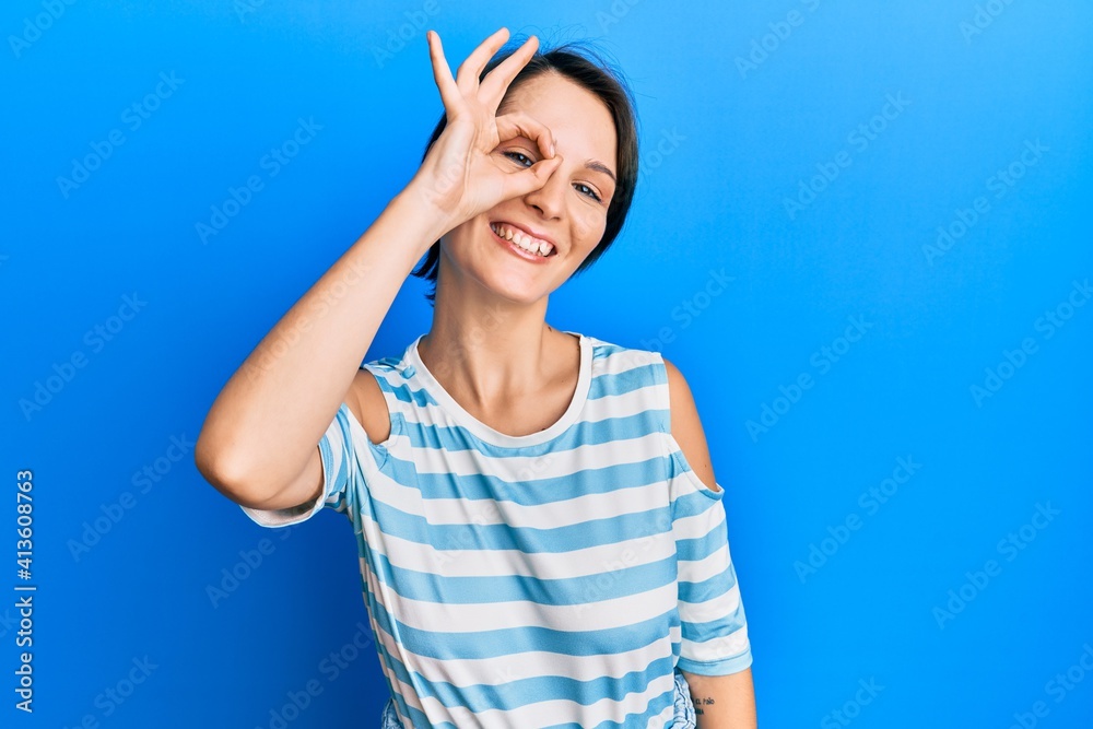 Young brunette woman with short hair wearing casual striped t-shirt smiling happy doing ok sign with hand on eye looking through fingers Young brunette woman with short hair wearing casual striped t-shirt smiling happy doing ok sign with hand on eye looking through fingers