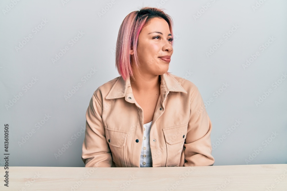 Hispanic woman with pink hair wearing casual clothes sitting on the table looking to side, relax profile pose with natural face and confident smile. Hispanic woman with pink hair wearing casual clothes sitting on the table looking to side, relax profile pose with natural face and confident smile.