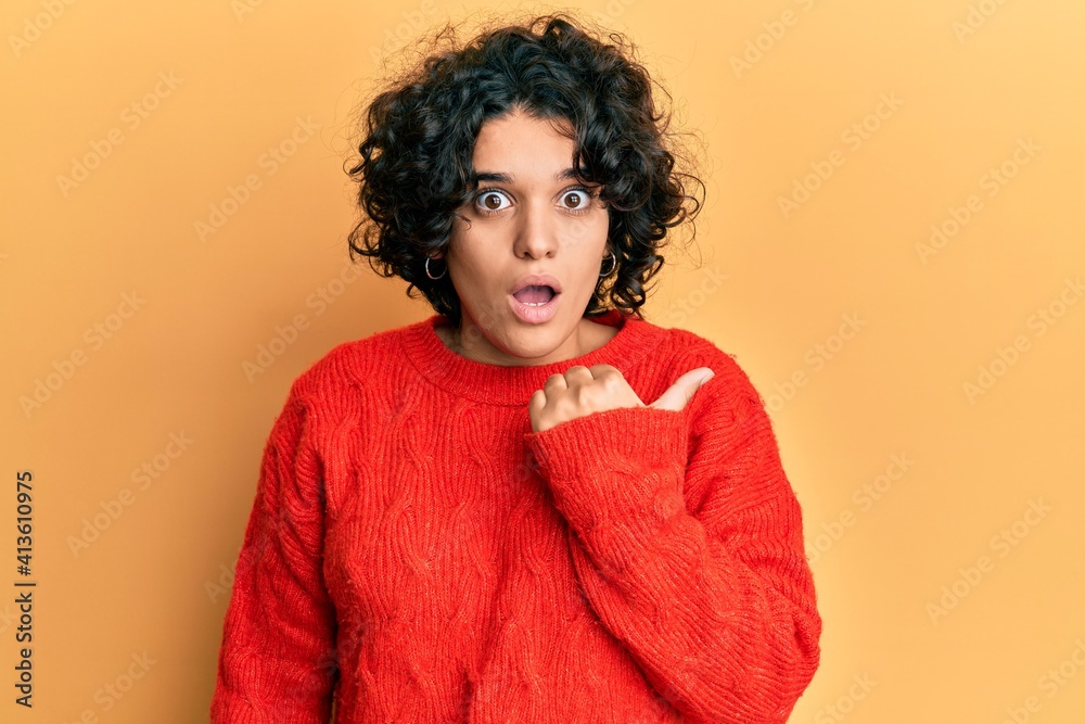 Young hispanic woman with curly hair wearing casual winter sweater surprised pointing with hand finger to the side, open mouth amazed expression. Young hispanic woman with curly hair wearing casual winter sweater surprised pointing with hand finger to the side, open mouth amazed expression.