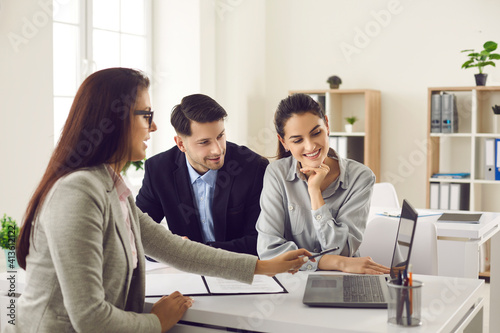 Happy future house buyers meeting real estate agent. Professional realtor talking to clients and offering flats options on computer. Smiling couple consulting bank worker or loan broker at her office