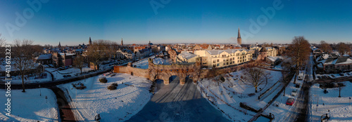Wide aerial panorama of Hanseatic medieval boat portal Berkelpoort and city wall of Zutphen with frozen over river and winter cityscape with snow  lit up by morning sun against a clear blue sky