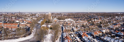 Winter aerial panorama of snow along the frozen canal going through the Dutch city of Zutphen with shadows of barren trees and former water tower in the background