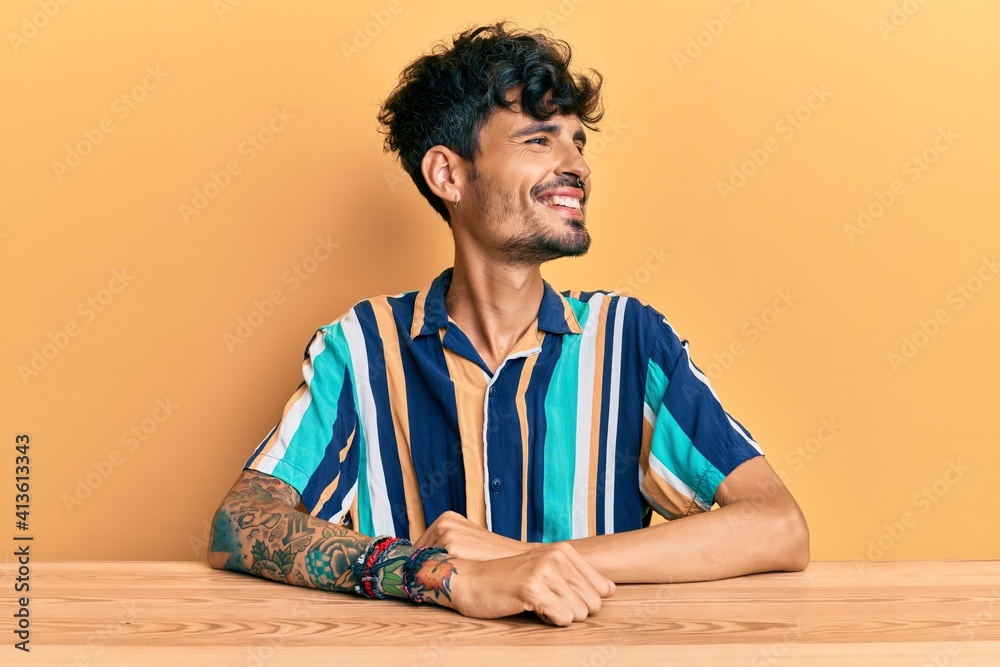 Young hispanic man wearing casual clothes sitting on the table looking away to side with smile on face, natural expression. laughing confident. Young hispanic man wearing casual clothes sitting on the table looking away to side with smile on face, natural expression. laughing confident.