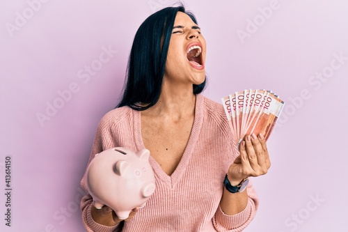 Young caucasian woman holding norwegian krona banknotes and piggy bank angry and mad screaming frustrated and furious, shouting with anger looking up. photo