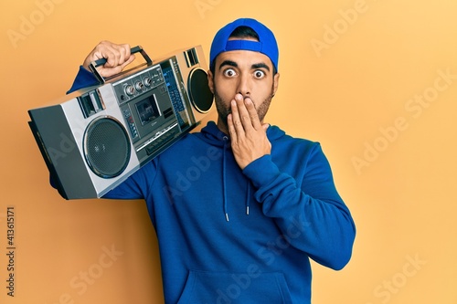 Young hispanic man holding boombox, listening to music covering mouth with hand, shocked and afraid for mistake. surprised expression
