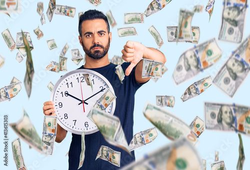 Young hispanic man holding big clock with angry face, negative sign showing dislike with thumbs down, rejection concept photo