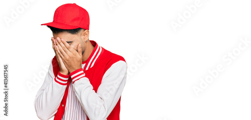 Young man with beard wearing baseball uniform with sad expression covering face with hands while crying. depression concept.