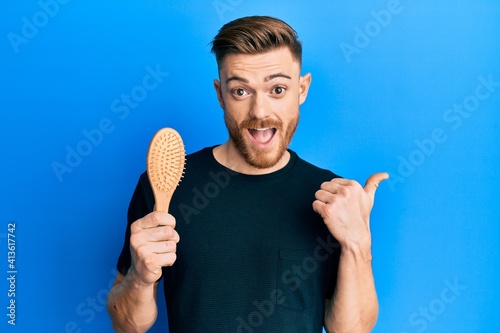 Young redhead man holding hairbrush pointing thumb up to the side smiling happy with open mouth