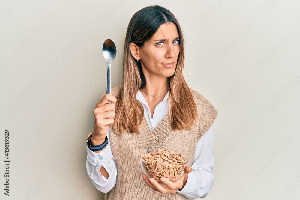 Brunette young woman eating healthy whole grain cereals with spoon clueless and confused expression. doubt concept. Brunette young woman eating healthy whole grain cereals with spoon clueless and confused expression. doubt concept.