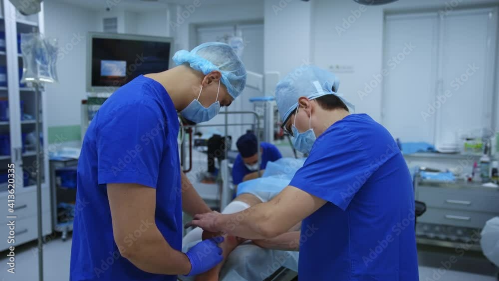Operation on leg. Medical specialists in blue uniform performing a surgery to a patient laying on surgical table in the modern operating theater. Operation on leg. Medical specialists in blue uniform performing a surgery to a patient laying on surgical table in the modern operating theater.