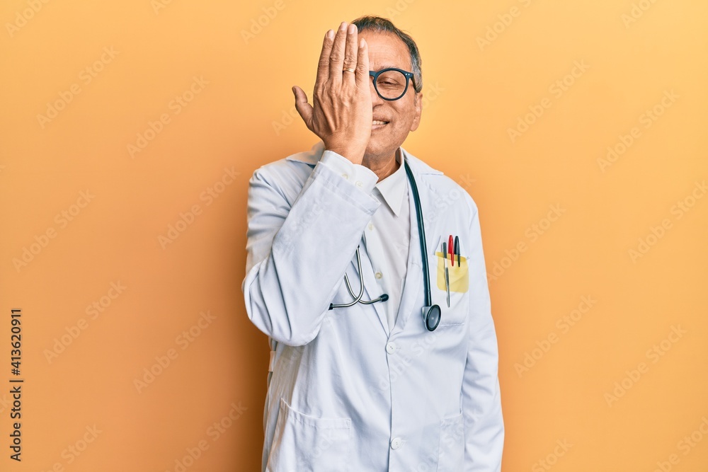 Middle age indian man wearing doctor coat and stethoscope covering one eye with hand, confident smile on face and surprise emotion. Middle age indian man wearing doctor coat and stethoscope covering one eye with hand, confident smile on face and surprise emotion.