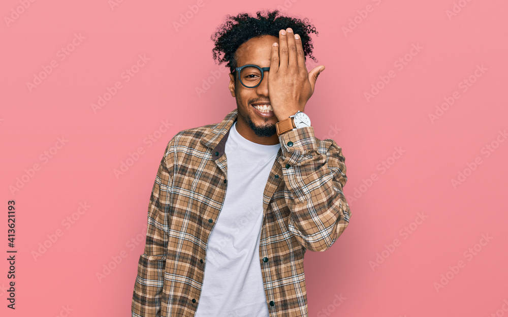 Young african american man with beard wearing casual clothes and glasses covering one eye with hand, confident smile on face and surprise emotion. Young african american man with beard wearing casual clothes and glasses covering one eye with hand, confident smile on face and surprise emotion.