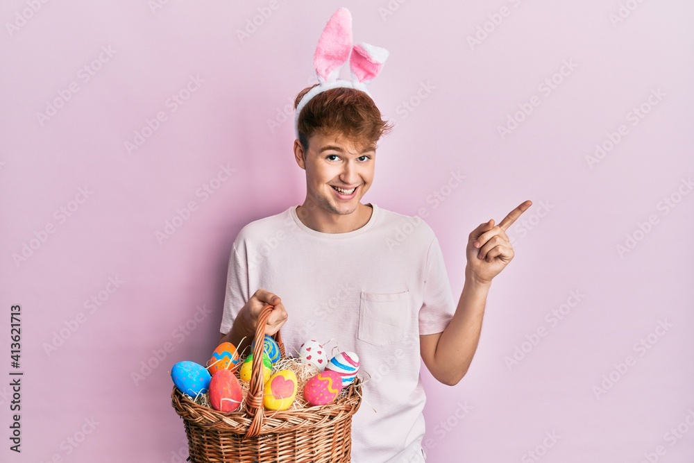 Young caucasian man wearing cute easter bunny ears holding wicker basket with colored eggs smiling happy pointing with hand and finger to the side