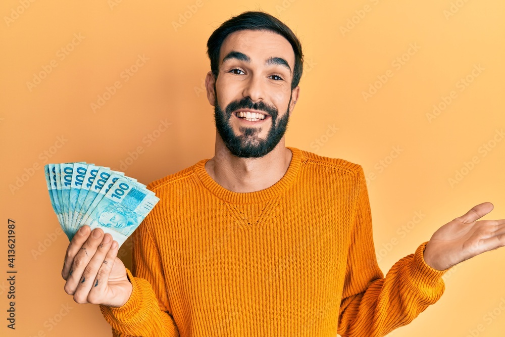 Young hispanic man holding 100 brazilian real banknotes celebrating achievement with happy smile and winner expression with raised hand Young hispanic man holding 100 brazilian real banknotes celebrating achievement with happy smile and winner expression with raised hand