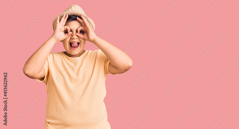 Little boy kid wearing summer hat and hawaiian swimsuit doing ok gesture like binoculars sticking tongue out, eyes looking through fingers. crazy expression. Little boy kid wearing summer hat and hawaiian swimsuit doing ok gesture like binoculars sticking tongue out, eyes looking through fingers. crazy expression.