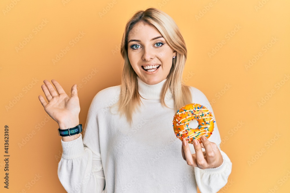 Young caucasian woman holding donut celebrating achievement with happy smile and winner expression with raised hand