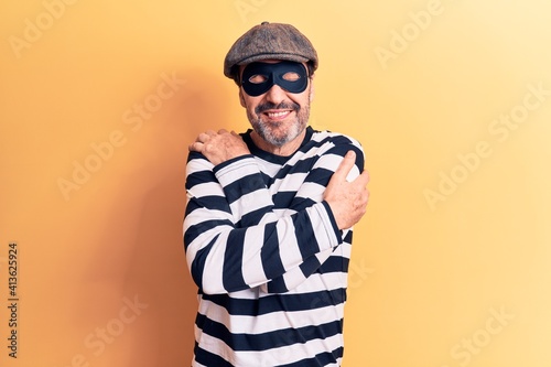 Middle age handsome burglar man wearing cap and mask over isolated yellow background hugging oneself happy and positive, smiling confident. Self love and self care