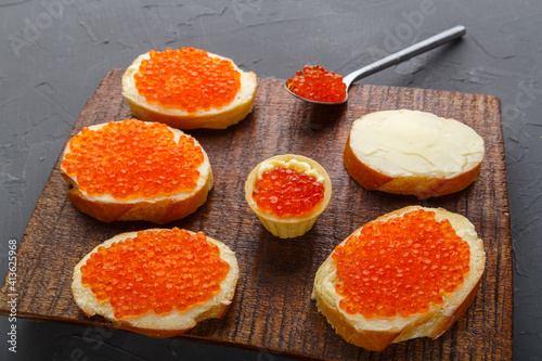 Bruschetta with butter and red caviar and a spoon with caviar on a brown board.