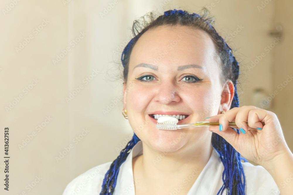 a woman in a white robe with a blue afro hairstyle in the bathroom near the mirror brushes her teeth face close.