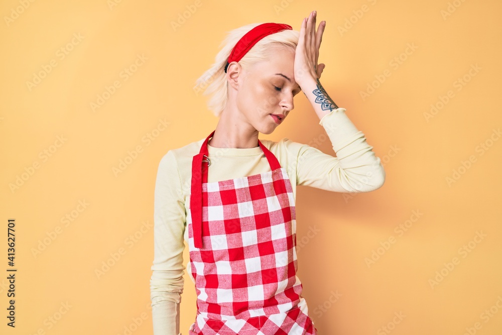 Young blonde woman with tattoo wearing professional baker apron surprised with hand on head for mistake, remember error. forgot, bad memory concept. Young blonde woman with tattoo wearing professional baker apron surprised with hand on head for mistake, remember error. forgot, bad memory concept.