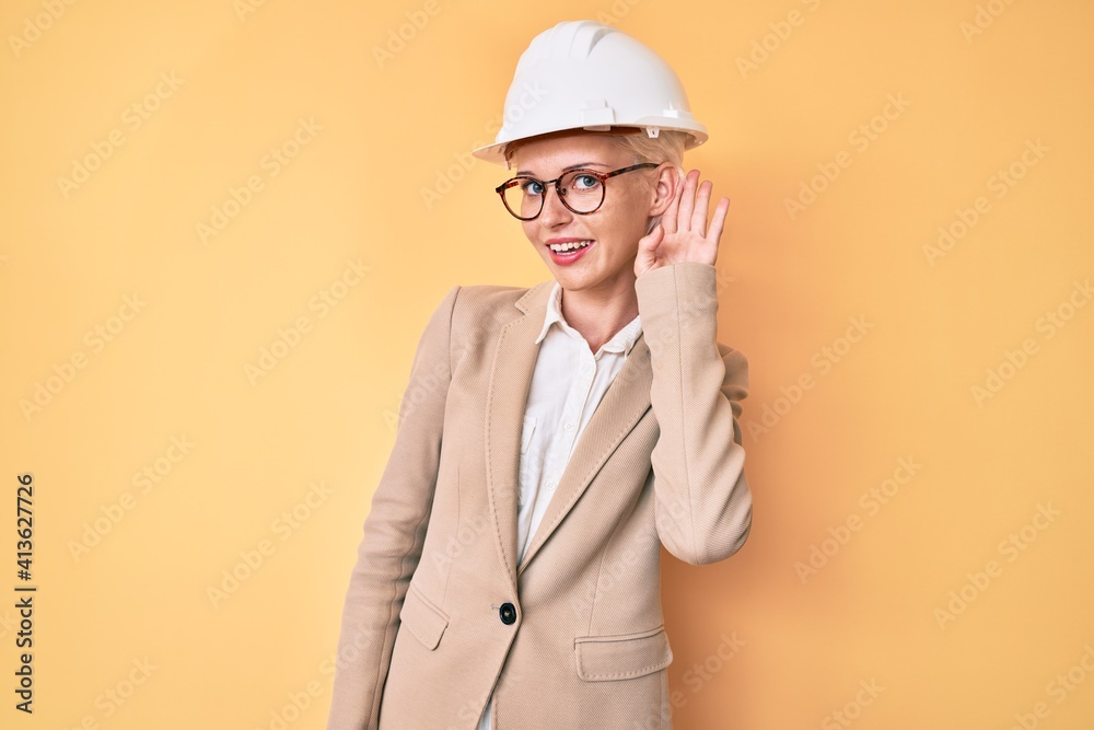 Young blonde woman wearing architect hardhat smiling with hand over ear listening and hearing to rumor or gossip. deafness concept.