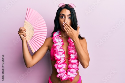 Beautiful hispanic woman wearing bikini holding handfan covering mouth with hand, shocked and afraid for mistake. surprised expression photo