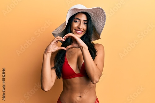 Beautiful hispanic woman wearing bikini and summer hat smiling in love doing heart symbol shape with hands. romantic concept.