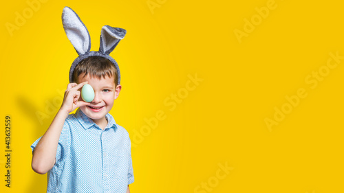 Happy child wearing bunny ears and holding colorful easter egg in front of his eye on a yellow background. Happy boy in easter bunny costume with easter egg