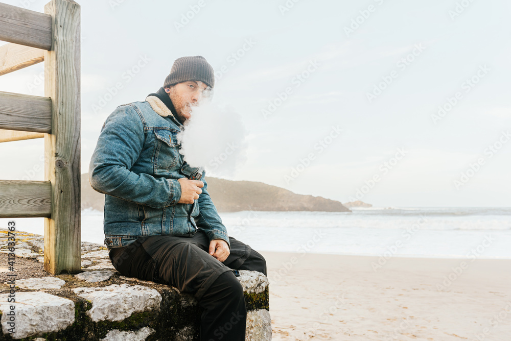 man vaping his electronic cigarette sitting on a wall in front of the beach, fuming and looking at the camera. alternative to tobacco. man vaping his electronic cigarette sitting on a wall in front of the beach, fuming and looking at the camera. alternative to tobacco.