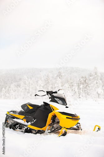 Yellow snowmobile is standing on a snowy field on a background of a winter scenic landscape.