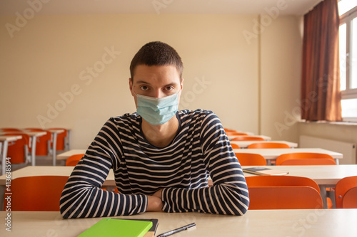 Young male student wearing a face mask and sitting at the desk in an empty classroom. Sad teenager missing his friends and classmates. Social distancing in school or university. New normal. 