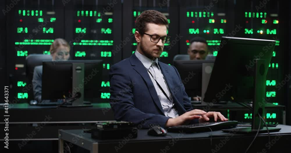 Senior IT worker with glasses sits in server room behind workplace and works with database on computer. Senior IT worker with glasses sits in server room behind workplace and works with database on computer.
