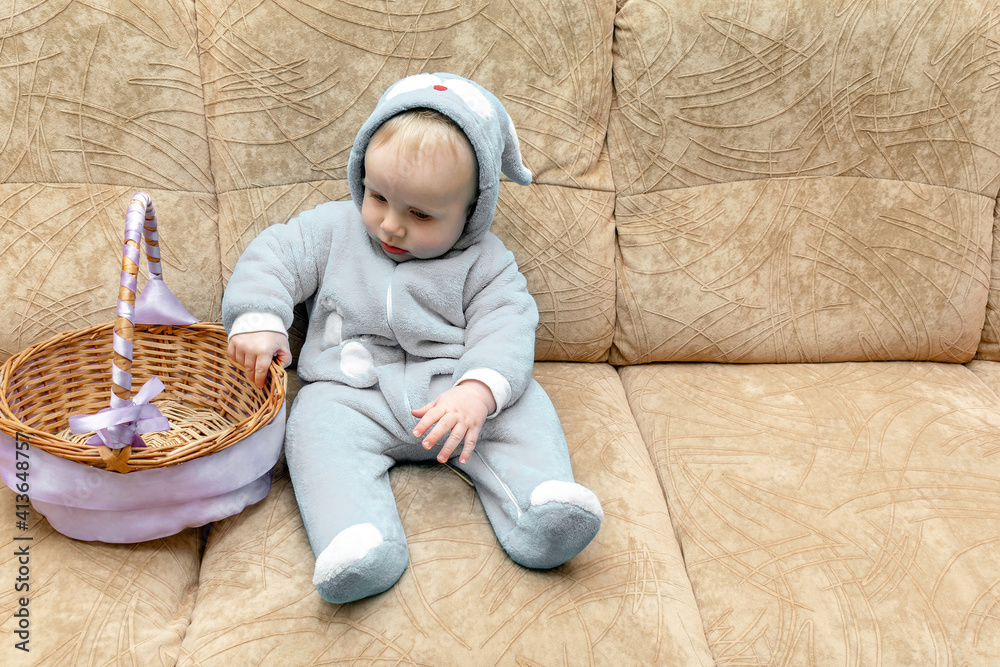 Naklejka premium Cute little toddler baby boy wearing a bunny, rabbit costume with basket sitting on couch. easter card