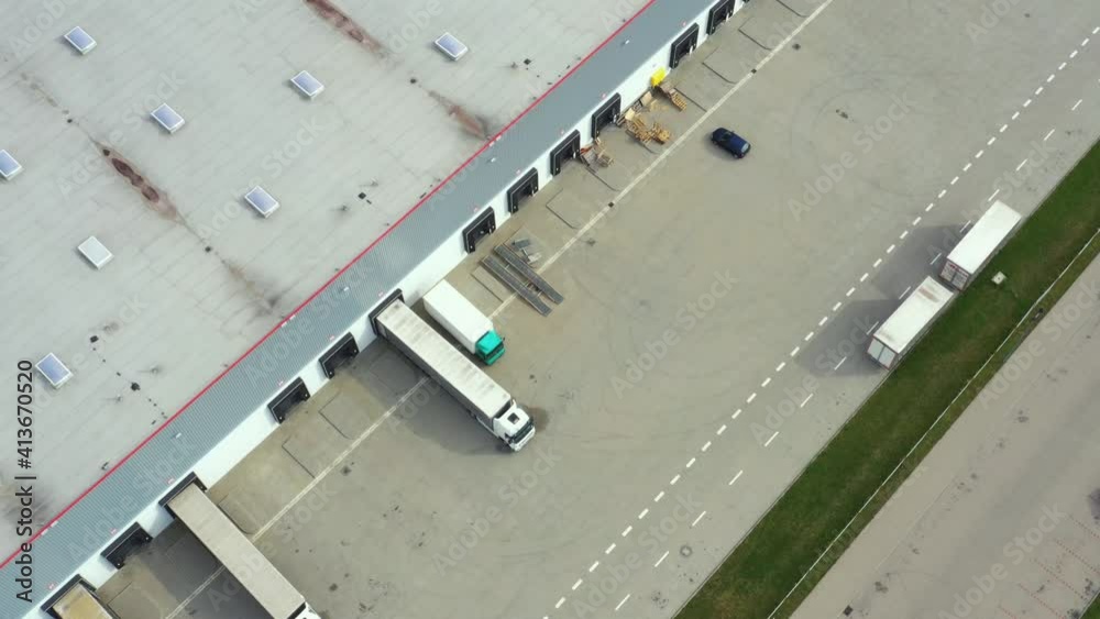 Aerial following shot of semi-trailer trucks travelling along a warehouse on the parking lot in the logistics park with loading hub. Aerial following shot of semi-trailer trucks travelling along a warehouse on the parking lot in the logistics park with loading hub.