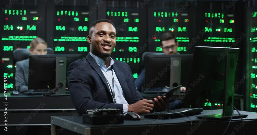 Smart African American IT worker sitting in server room behind workplace holding tablet working with database raises his head and looks at camera smiling. Smart African American IT worker sitting in server room behind workplace holding tablet working with database raises his head and looks at camera smiling.