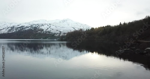 White snow covered the tops of the mountain in the background with trees in autumn colors next to loch Lomond in Scotland. Drone trucking shot photo