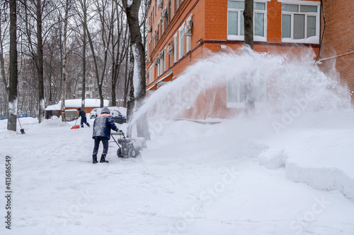 A utility worker cleans a path with a snow blower during heavy snow on a winter day. A jet of snow takes off into the air.