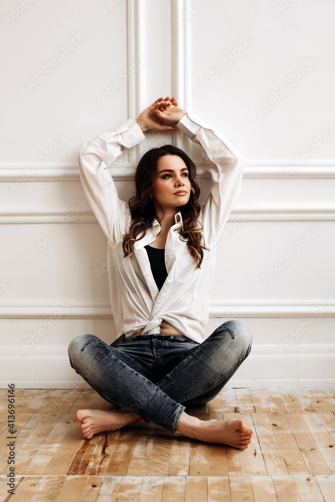 A beautiful model sits on the floor in a countless interior. happy young woman in a white shirt and jeans raises her hands up. 
