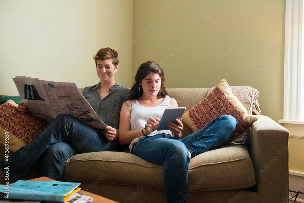Woman using tablet by girlfriend reading newspaper while sitting on couch at home Woman using tablet by girlfriend reading newspaper while sitting on couch at home