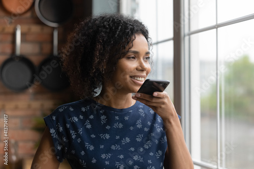 Close up smiling African American woman recording voice message, standing near window at home, holding smartphone, chatting online by speakerphone, activating digital assistant on smartphone