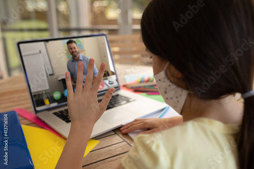 Fototapeta Female student wearing face mask having a video call with male teacher on laptop at school