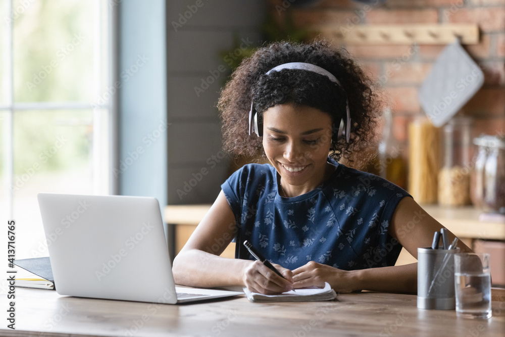 Close up smiling African American woman in headphones writing taking notes, using laptop, listening to online lecture, course, happy student studying at home, watching webinar, preparing to exam Close up smiling African American woman in headphones writing taking notes, using laptop, listening to online lecture, course, happy student studying at home, watching webinar, preparing to exam