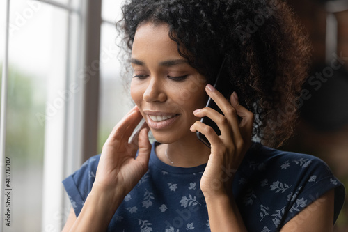 Close up smiling African American woman talking on phone, enjoying pleasant conversation, attractive young female holding smartphone, chatting with friend or boyfriend, standing near window at home