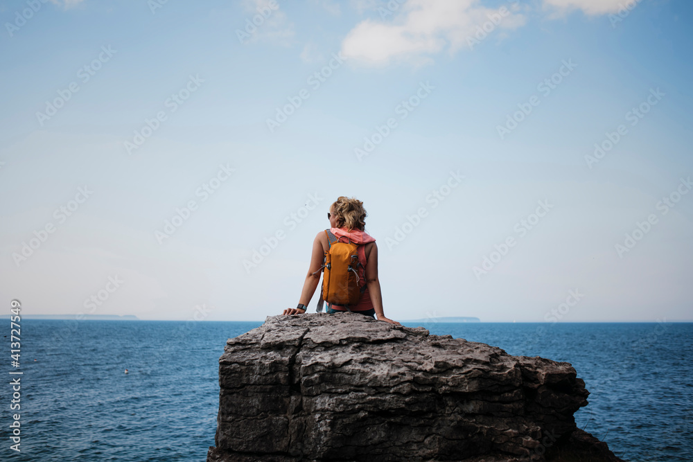 Rear view of female hiker with backpack sitting on rock against sea and sky at Bruce Peninsula National Park Rear view of female hiker with backpack sitting on rock against sea and sky at Bruce Peninsula National Park