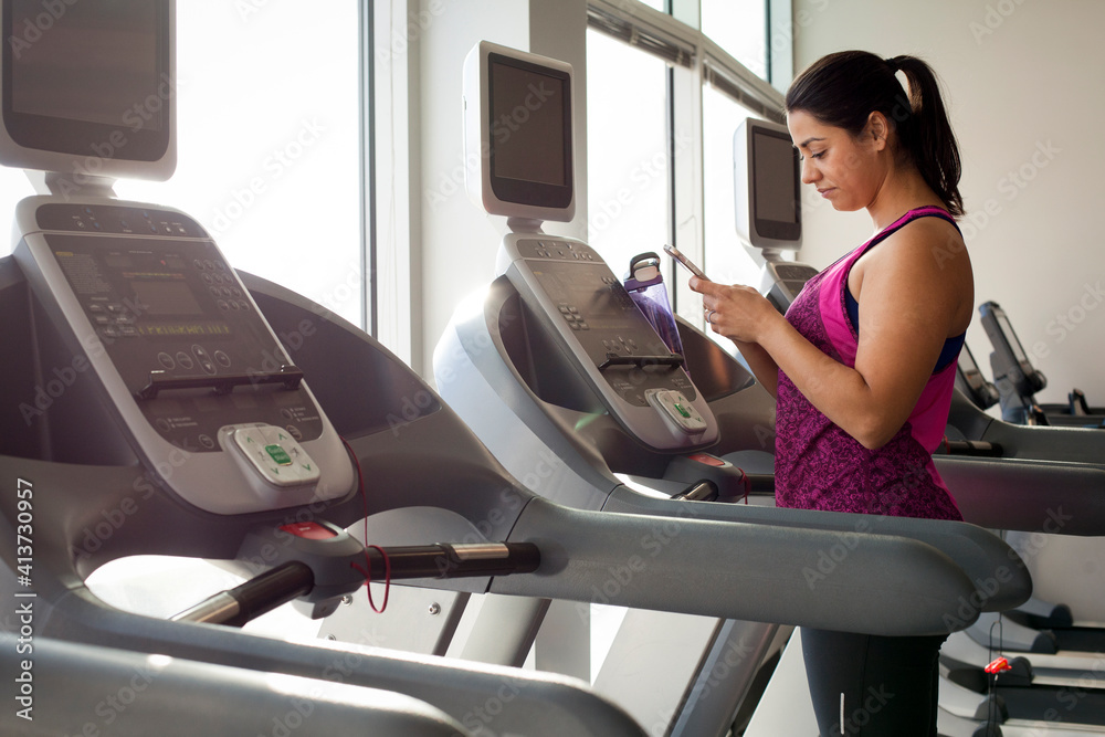 Side view of woman using smart phone while standing on treadmill in gym Side view of woman using smart phone while standing on treadmill in gym